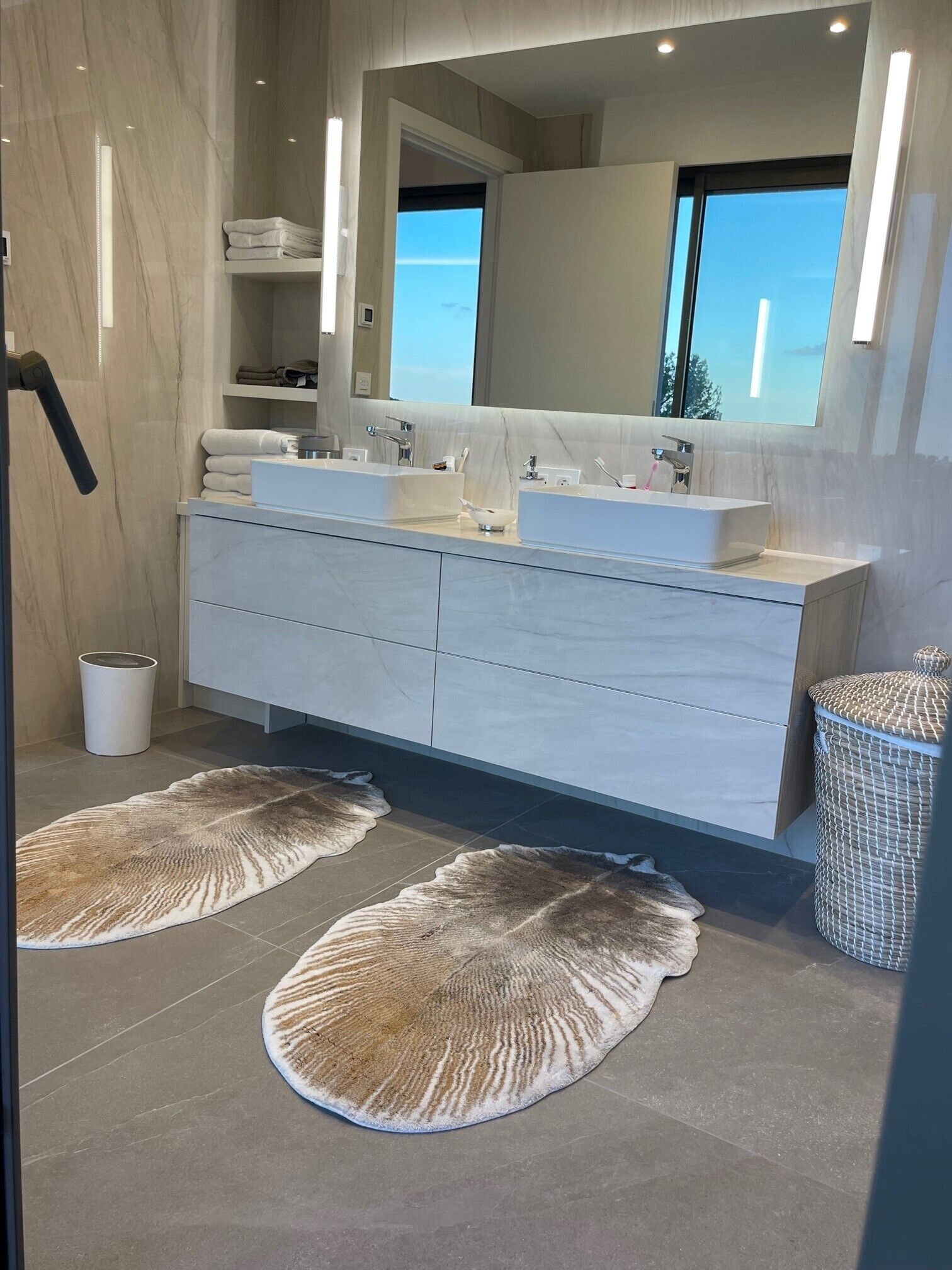 A modern bathroom with two sinks, a large mirror, and a view of the outdoors.