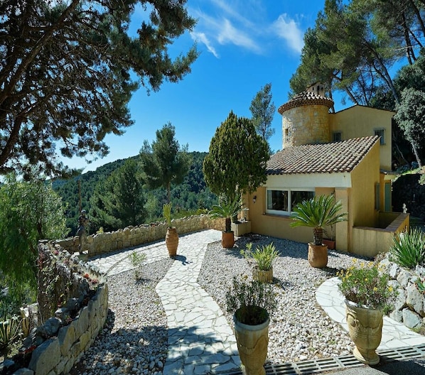 Eine mediterrane Villa mit einem steinbelagten Weg, der zu einem gelben Gebäude führt, umgeben von Kübelpflanzen und Bäumen.