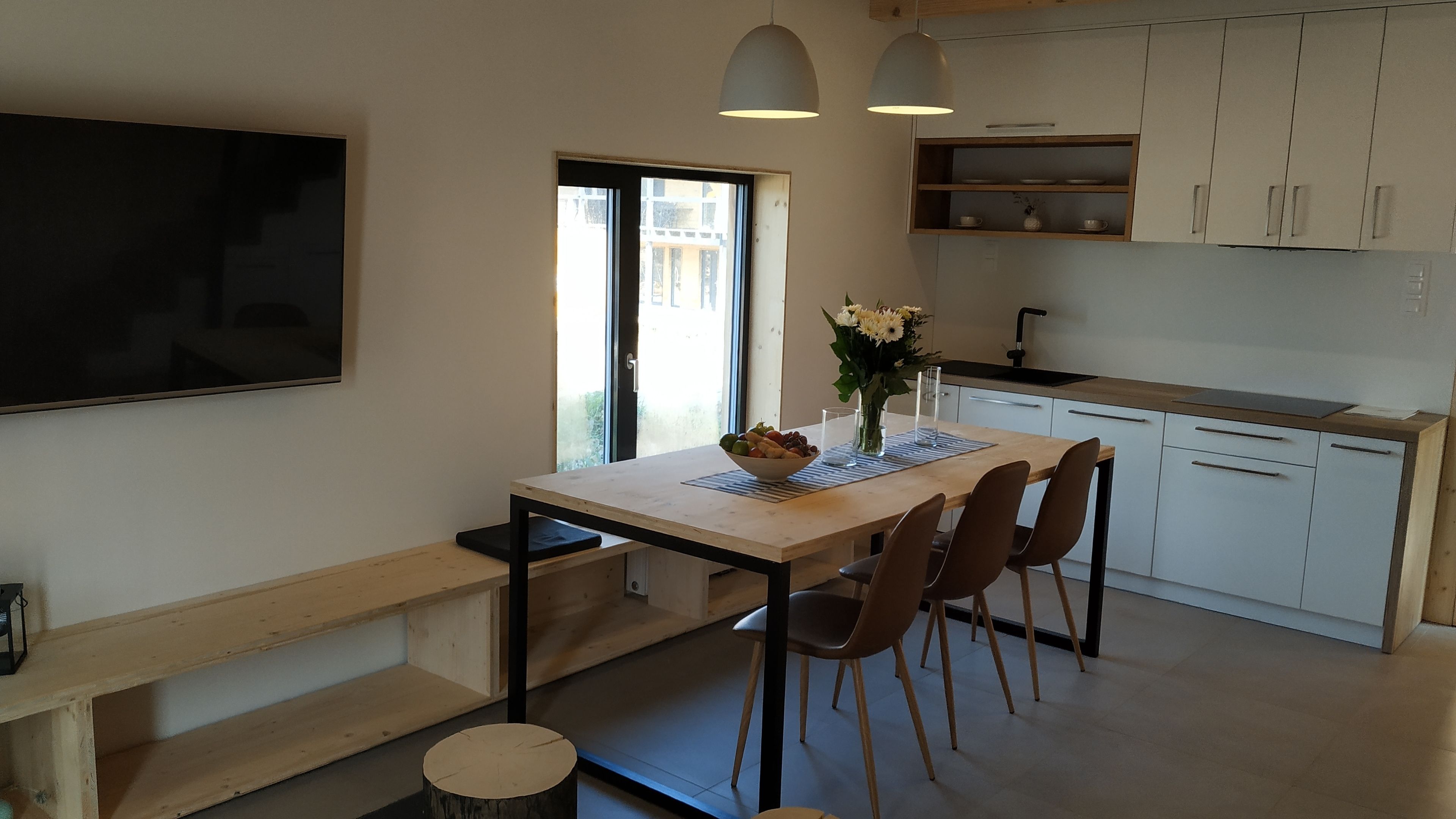 A modern kitchen with a dining area, featuring a large window, wooden cabinets, and a flat-screen TV mounted on the wall.