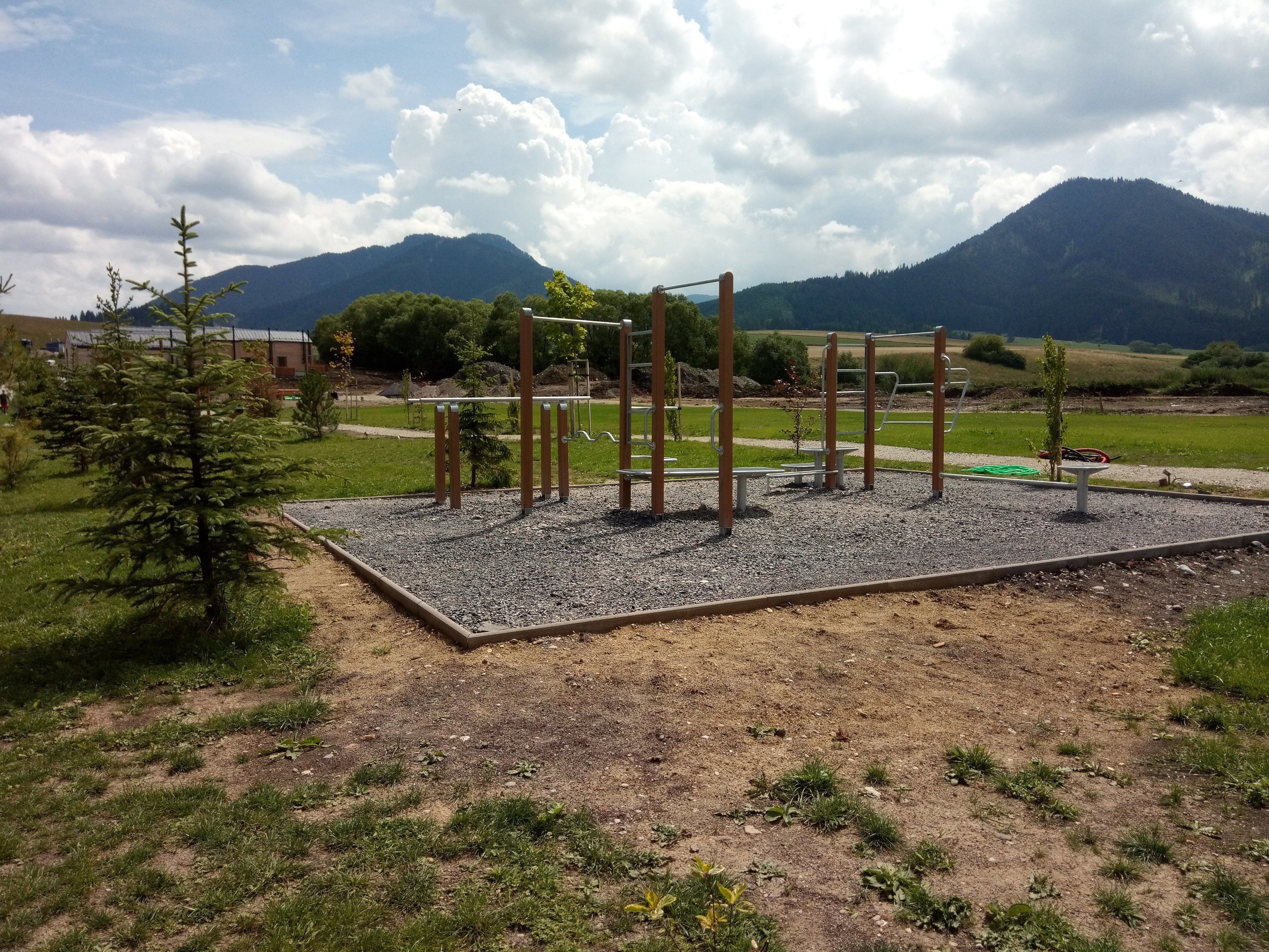Un parque infantil con equipamiento de ejercicio, rodeado de grava y césped, con montañas al fondo.