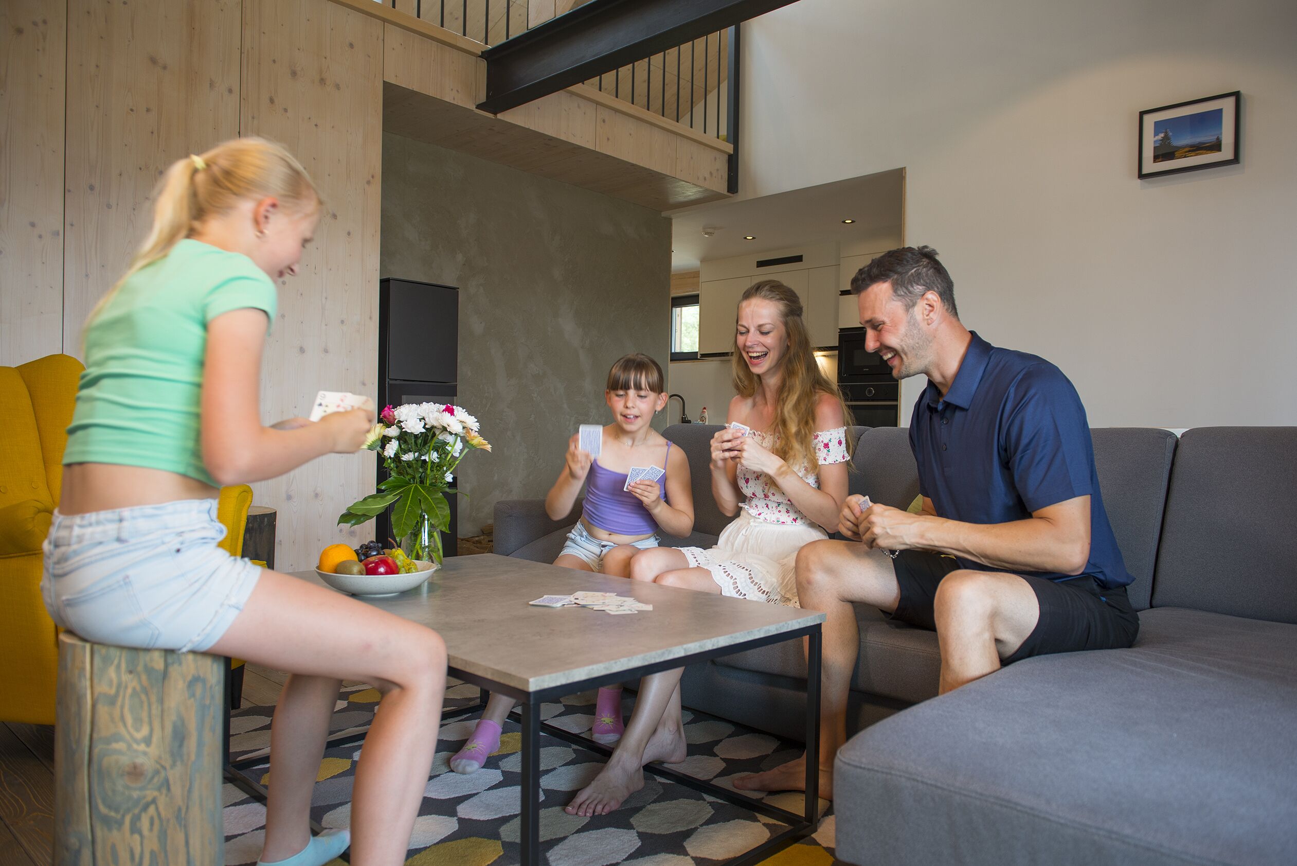 Une famille de quatre personnes est assise autour d’une table basse dans un salon moderne aux couleurs neutres, agrémenté d’éléments en bois et doté d’un design contemporain.