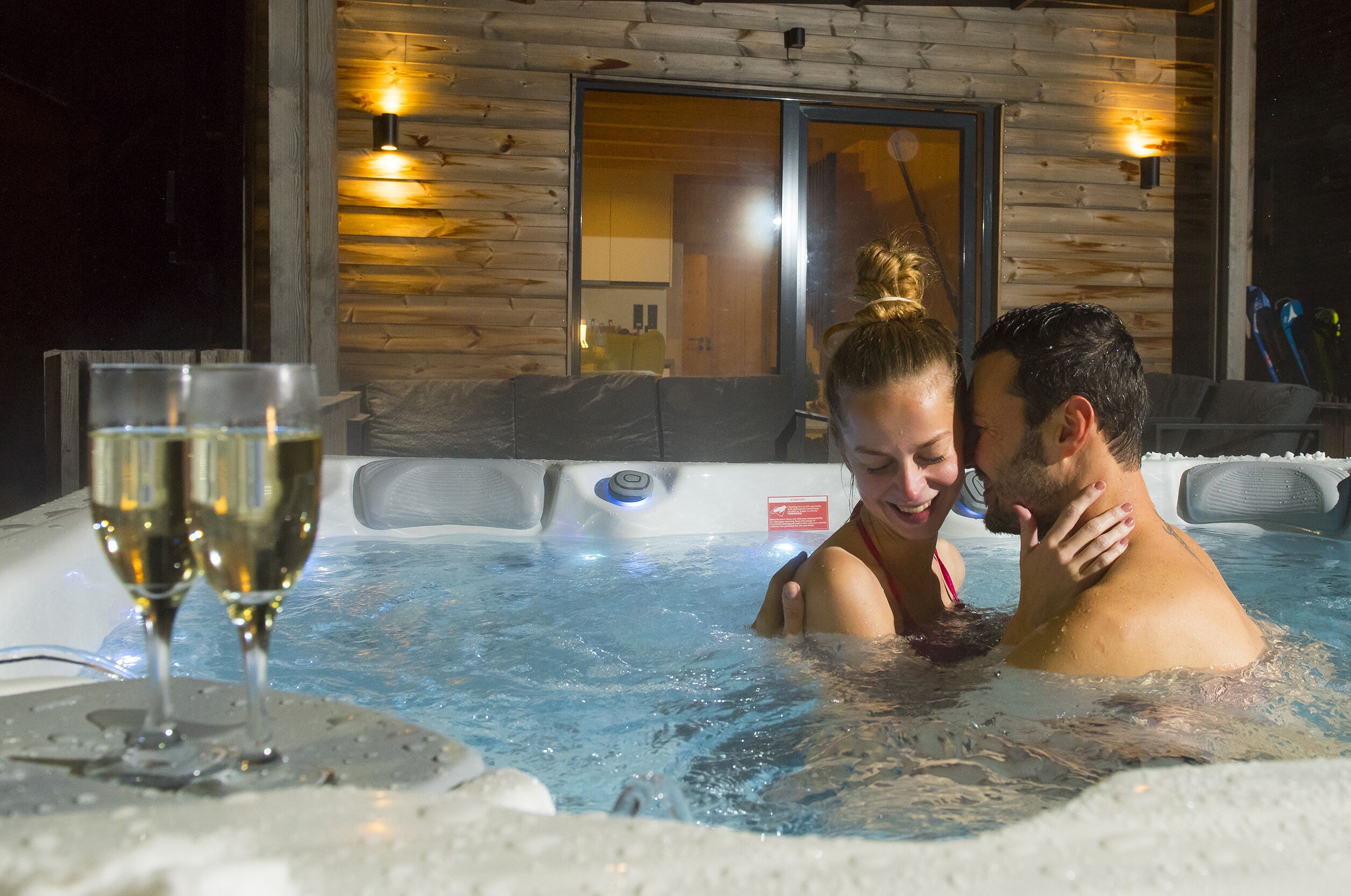 A couple in a hot tub with a glass of champagne, wooden cabin-style interior, and a view of the interior through a glass door.