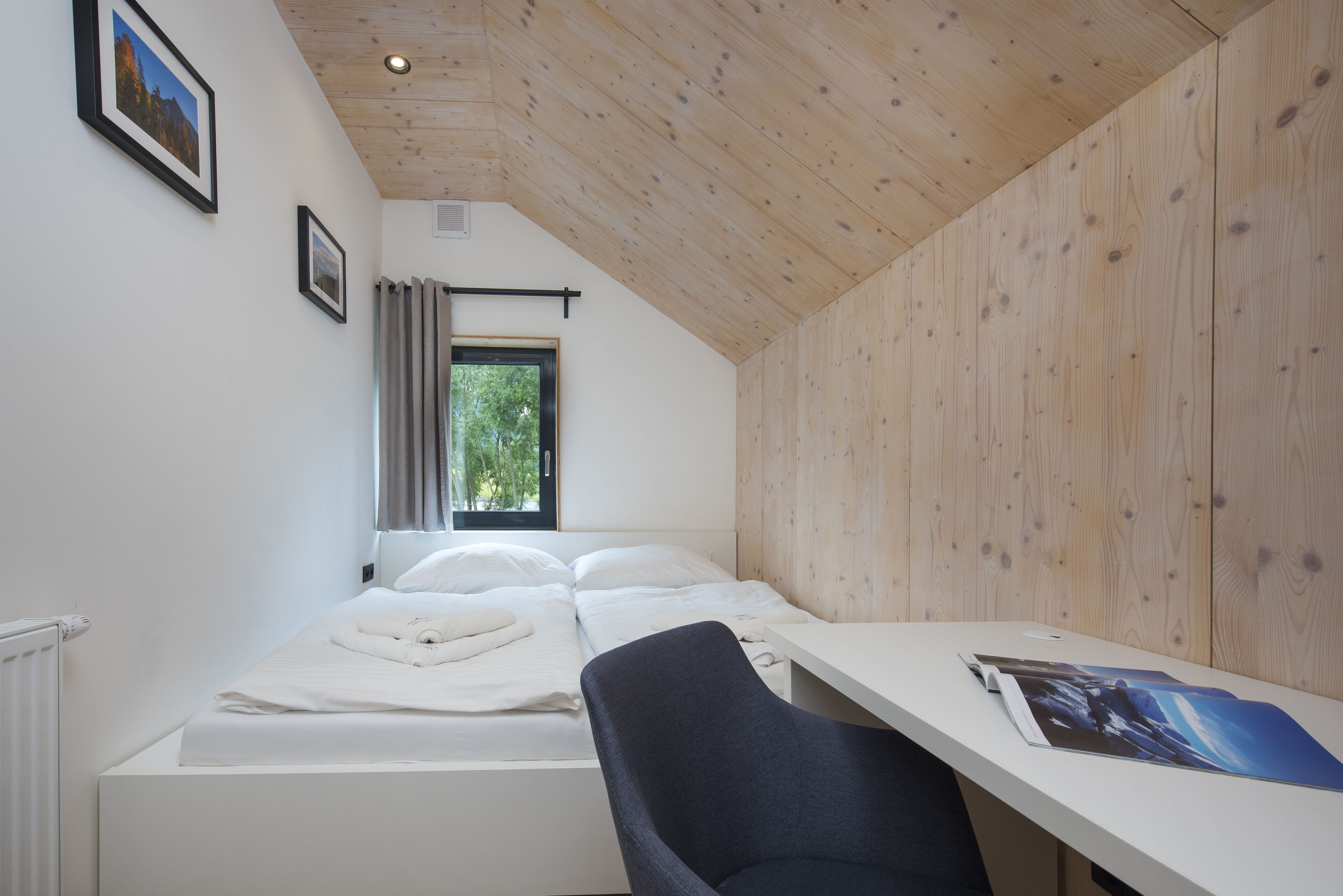Un dormitorio con techo de madera, cama con ropa de cama blanca, escritorio con silla y ventana con cortinas.