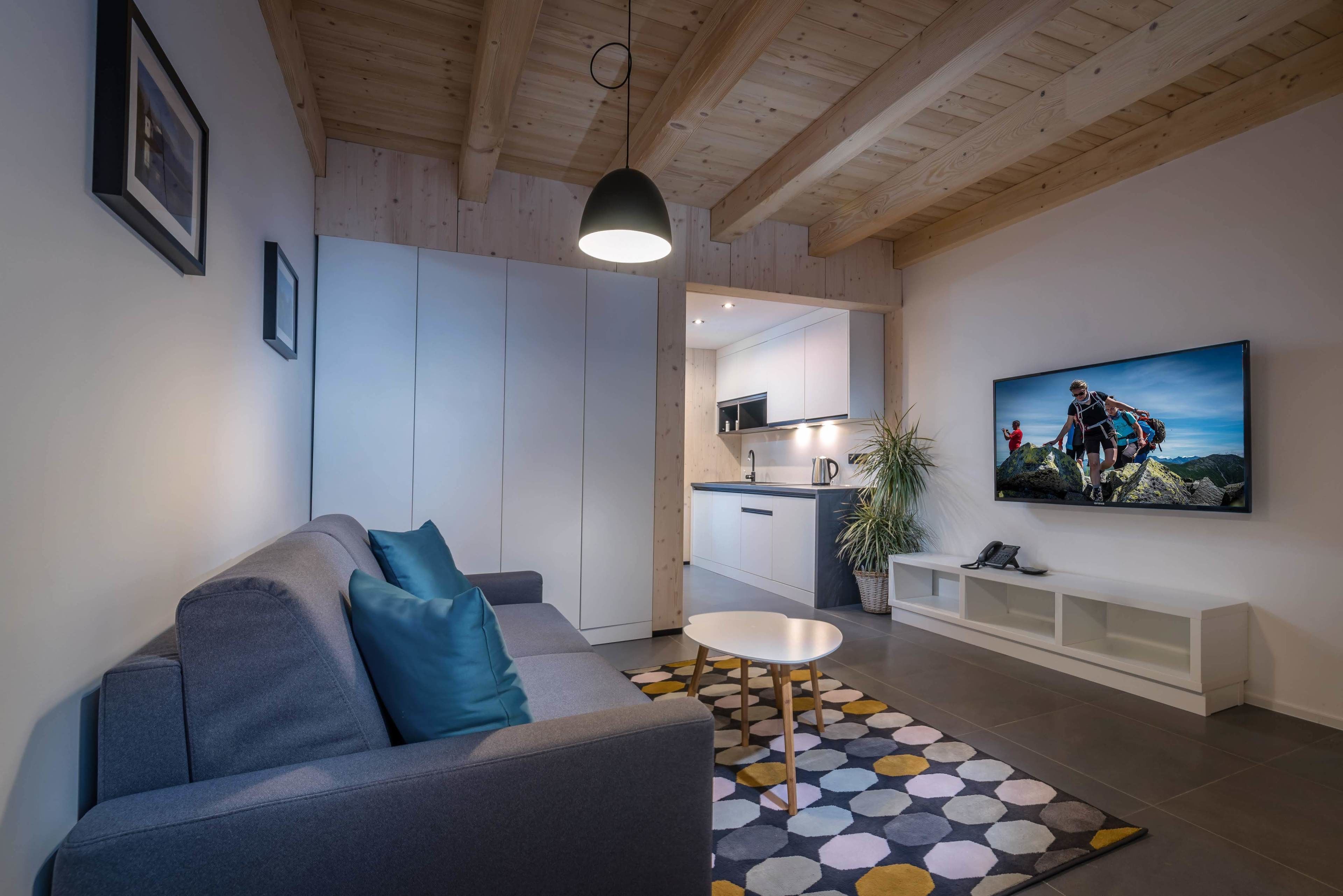 Una sala de estar moderna con un sofá gris, una mesa de centro blanca y una alfombra colorida. Un televisor está montado en la pared, y se aprecia una cocina al fondo.