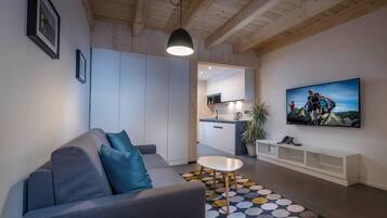 Una sala de estar moderna con un sofá gris, una mesa de centro blanca y una alfombra colorida. Un televisor está montado en la pared, y se aprecia una cocina al fondo.