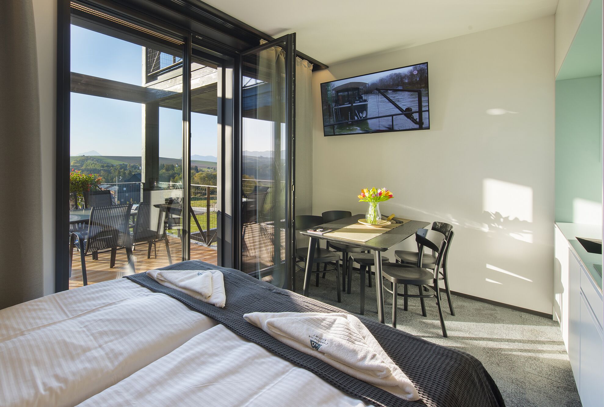 A hotel room with a balcony, a dining table with chairs, a TV mounted on the wall, and a bed with a view of the outdoors.