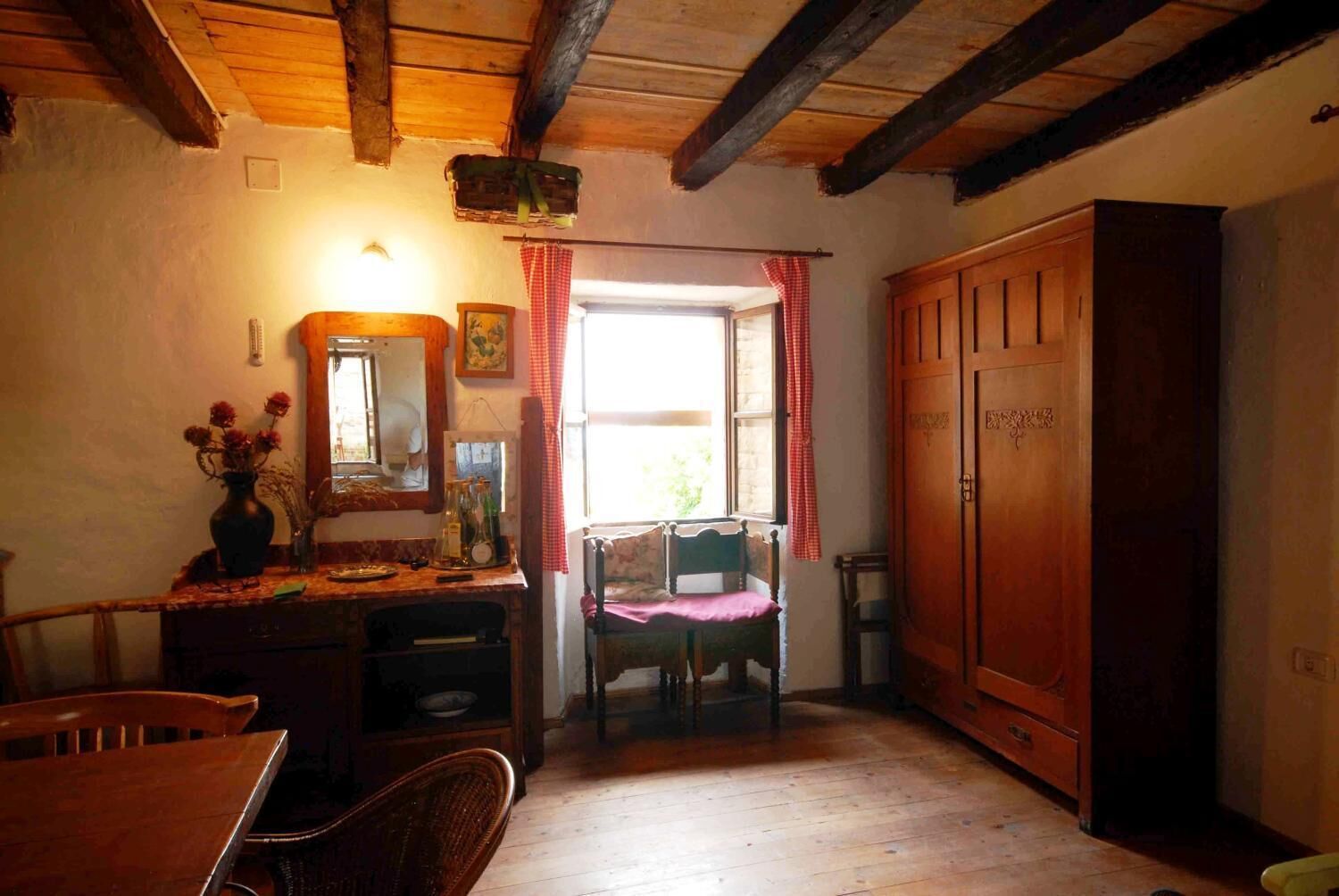 A rustic room with a wooden ceiling, a large wooden wardrobe, a chair with a pink cushion, a wooden table with a vase of flowers, and a window with red curtains.