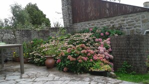 A stone wall with a garden of pink and white flowers, a table, and a stone floor. - Very nice renovated family house (DINAN)