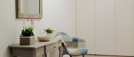 A room with a white wardrobe, a mirror, a table with two potted plants, and a chair.