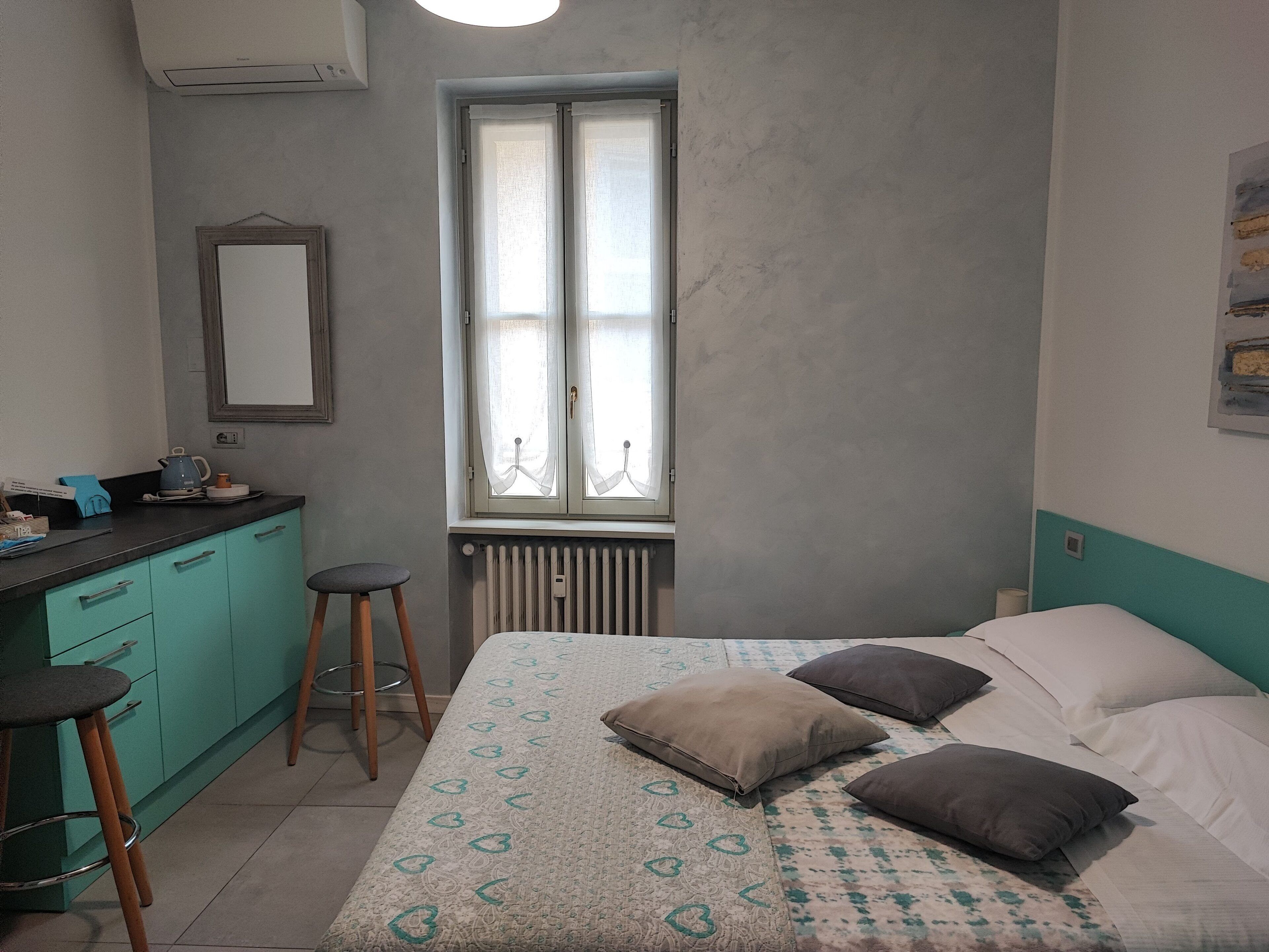 Una camera da letto con un letto, una finestra, un termosifone, uno specchio e un angolo cottura.