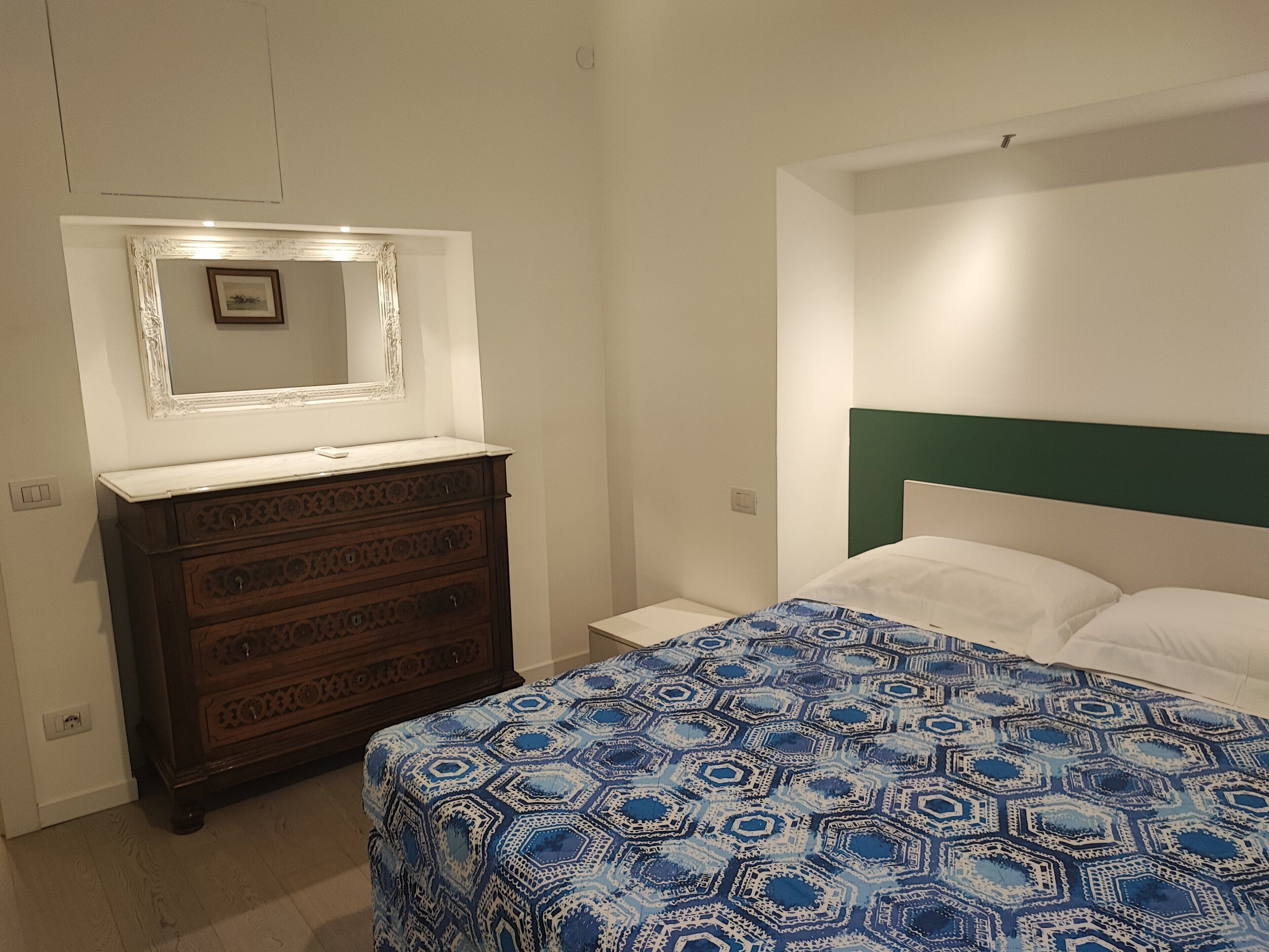A bedroom with a wooden dresser, a bed with a patterned bedspread, a nightstand, and a mirror above the dresser.