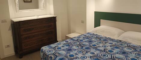A bedroom with a wooden dresser, a bed with a patterned bedspread, a nightstand, and a mirror above the dresser.