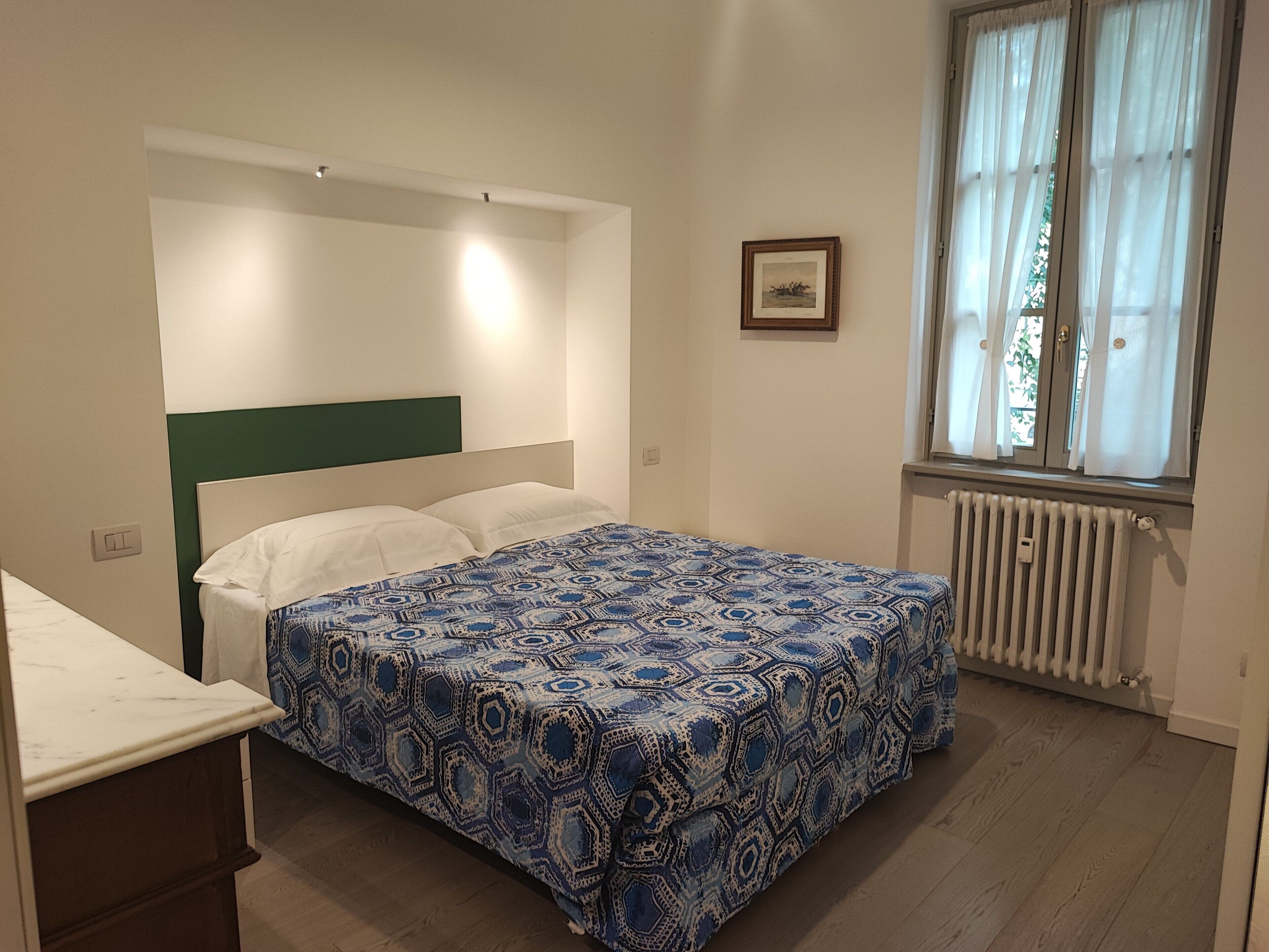 A bedroom with a bed, a window with curtains, a radiator, and a framed picture on the wall.