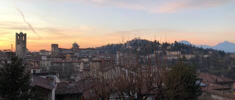 A town with historic architecture, including a prominent tower and a church with a dome, set against a backdrop of mountains and a sunset sky.