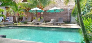 A swimming pool with lounge chairs and umbrellas, surrounded by tropical plants and a thatched-roof structure.