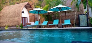 A poolside area with lounge chairs and umbrellas, a thatched-roof building, and a stone wall.