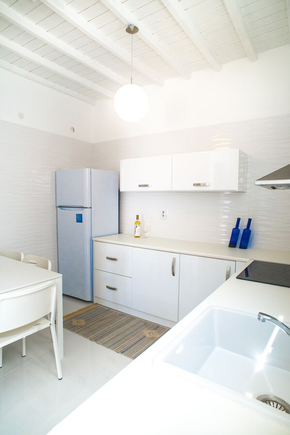 A modern kitchen with white cabinetry, a built-in refrigerator, a countertop with a sink, and a dining area with a table and chairs. - Chora House (Mykonos)