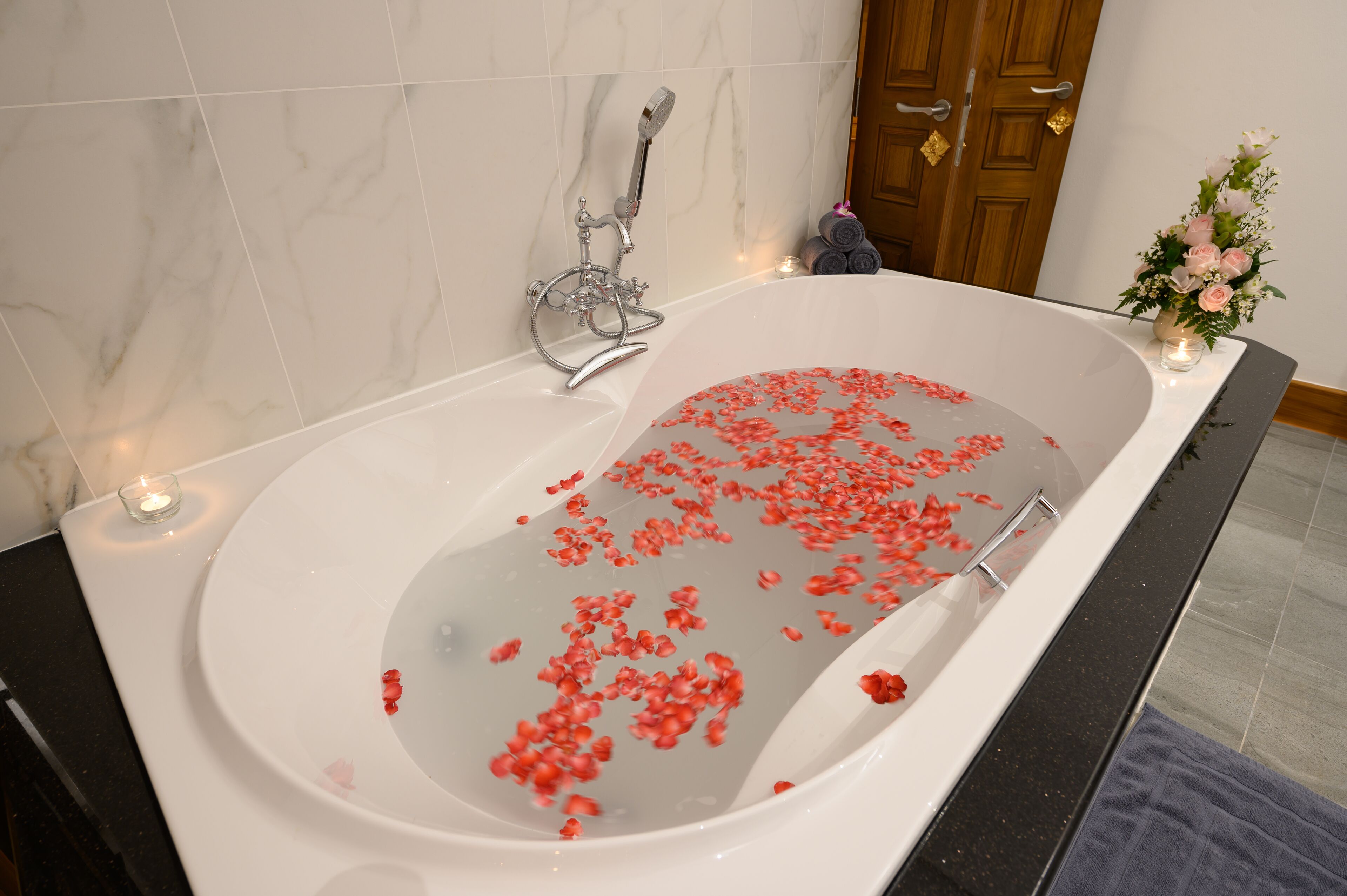 A bathtub filled with water and rose petals, a showerhead, and a small candle on the side.