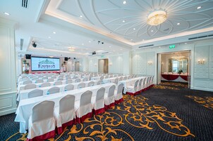 A conference room with rows of tables, white chairs, and a large screen displaying the hotel name.