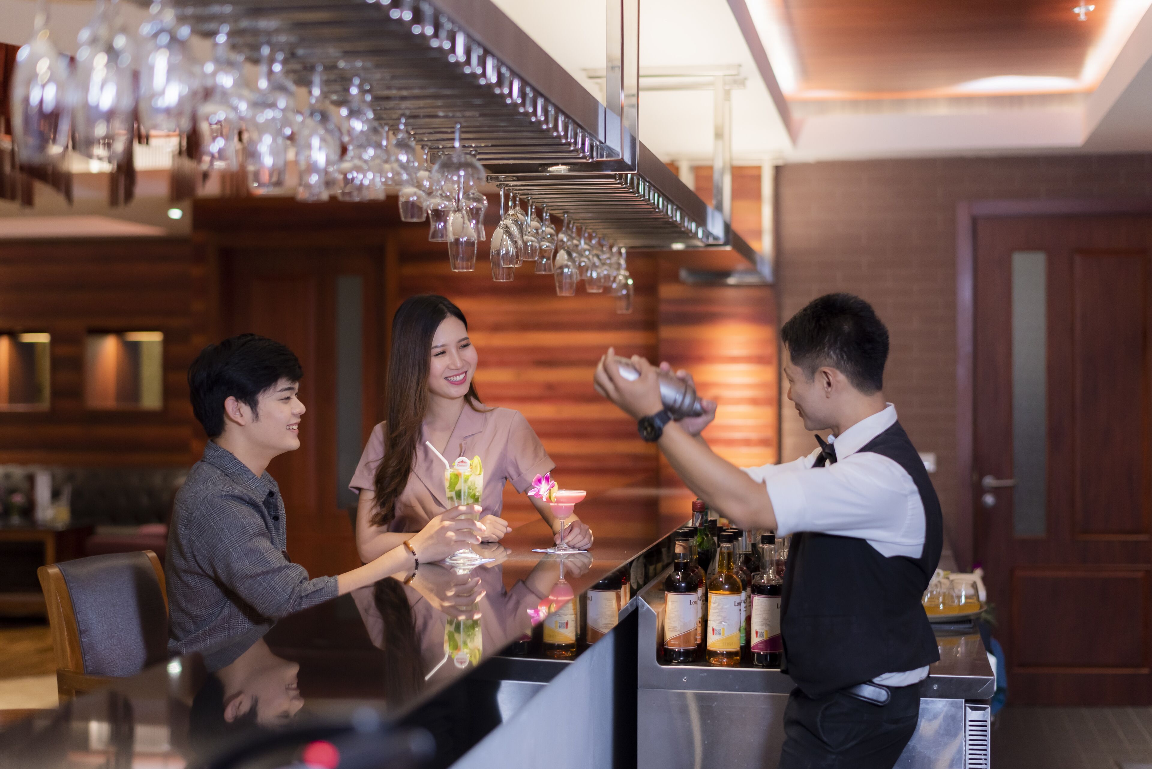 A bar area with a bartender preparing drinks, a woman holding a cocktail, and a man sitting at the bar.