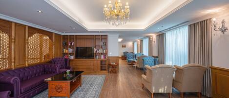 A spacious living room with a large chandelier, a purple sofa, beige armchairs, and a wooden bookshelf.