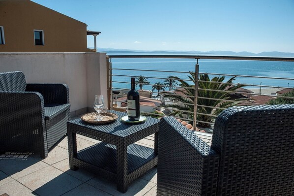 Un balcone con mobili in vimini, un bicchiere d'acqua e una bottiglia di vino, con vista sulla costa, palme e edifici.