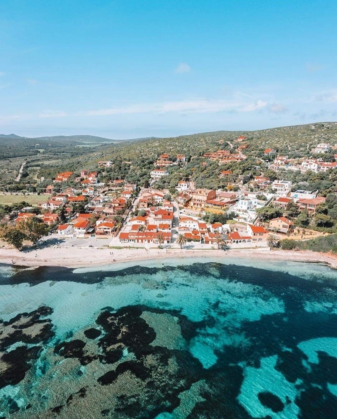Vue aérienne d’une ville côtière avec des bâtiments aux toits rouges, une plage et des eaux turquoise cristallines.