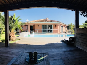 A covered outdoor area with a pool, wooden deck, and a house in the background. - DETACHED UPSTAIRS VILLA WITH PRIVATE POOL AND GARDEN (Porto Vecchio)