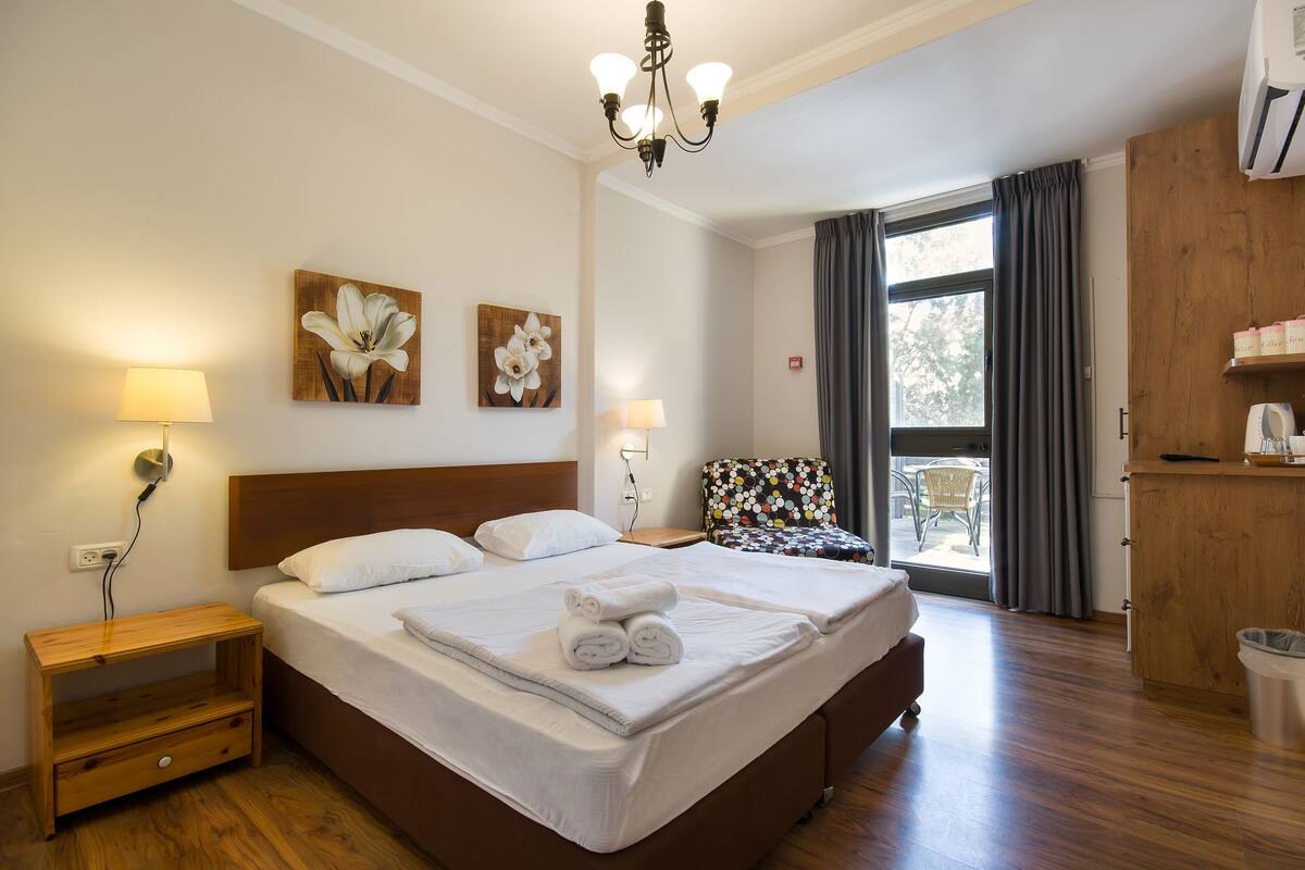 A hotel room with a large bed, two wall-mounted lamps, a wooden nightstand, a small table, and a view of the outdoors through a window.