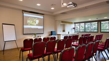 Une salle de conférence avec des chaises rouges, un tableau blanc, un écran de projection et une table sur laquelle se trouvent un ordinateur portable et une plante.
