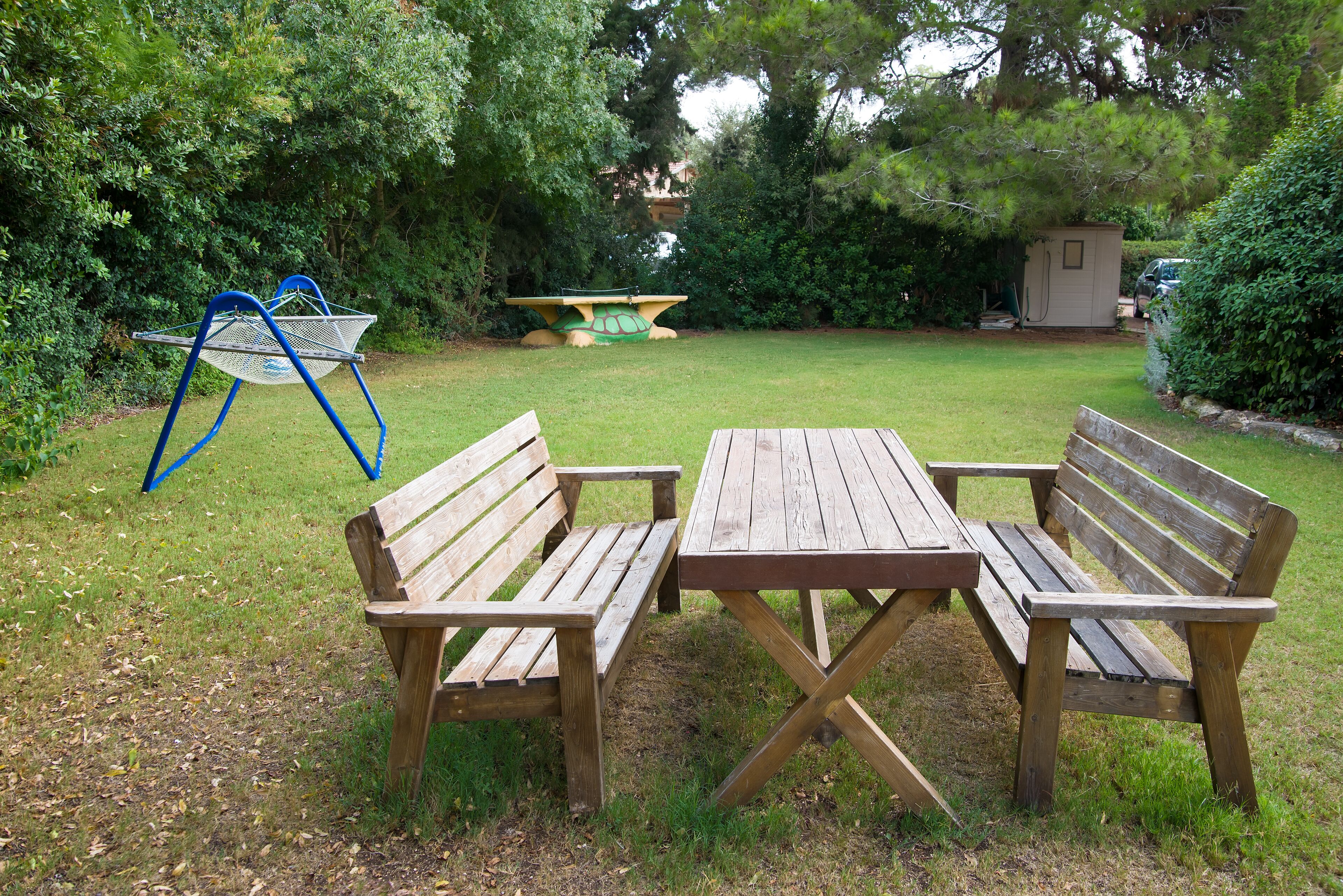 A wooden bench and table set in a grassy garden area with a metal chair and a small shed in the background.