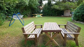 A wooden bench and table set in a grassy garden area with a metal chair and a small shed in the background.