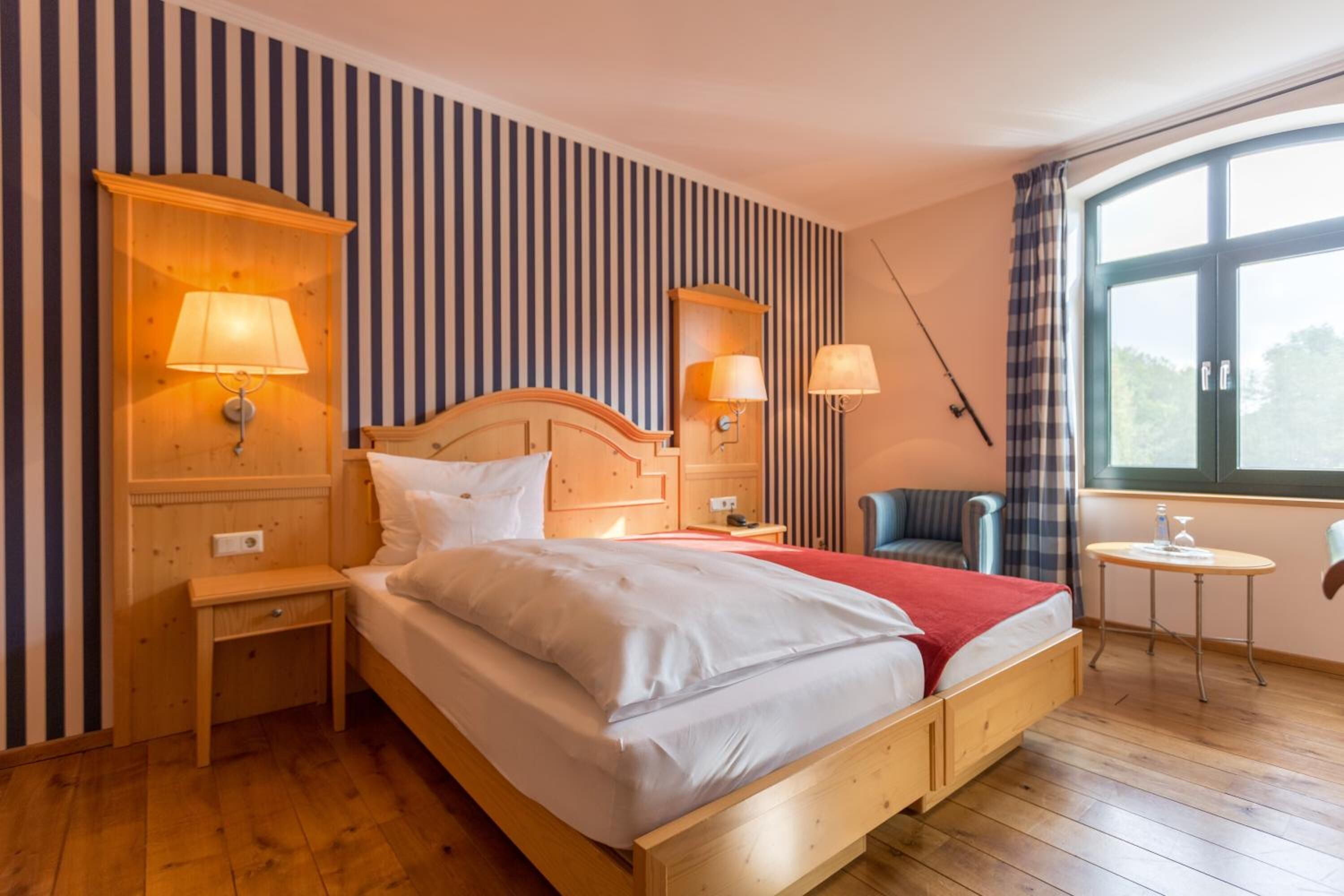 Una habitación de hotel con una cama de madera, dos mesitas de noche con lámparas, una silla, una mesa redonda pequeña y un ventanal con cortinas.