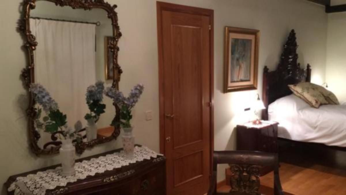 A bedroom with a wooden door, a large mirror, a bedside table with a lamp, and a bed with pillows.