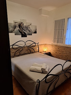 Une chambre à coucher avec un lit, une œuvre d’art accrochée au mur, une fenêtre avec des stores et une lampe de chevet.