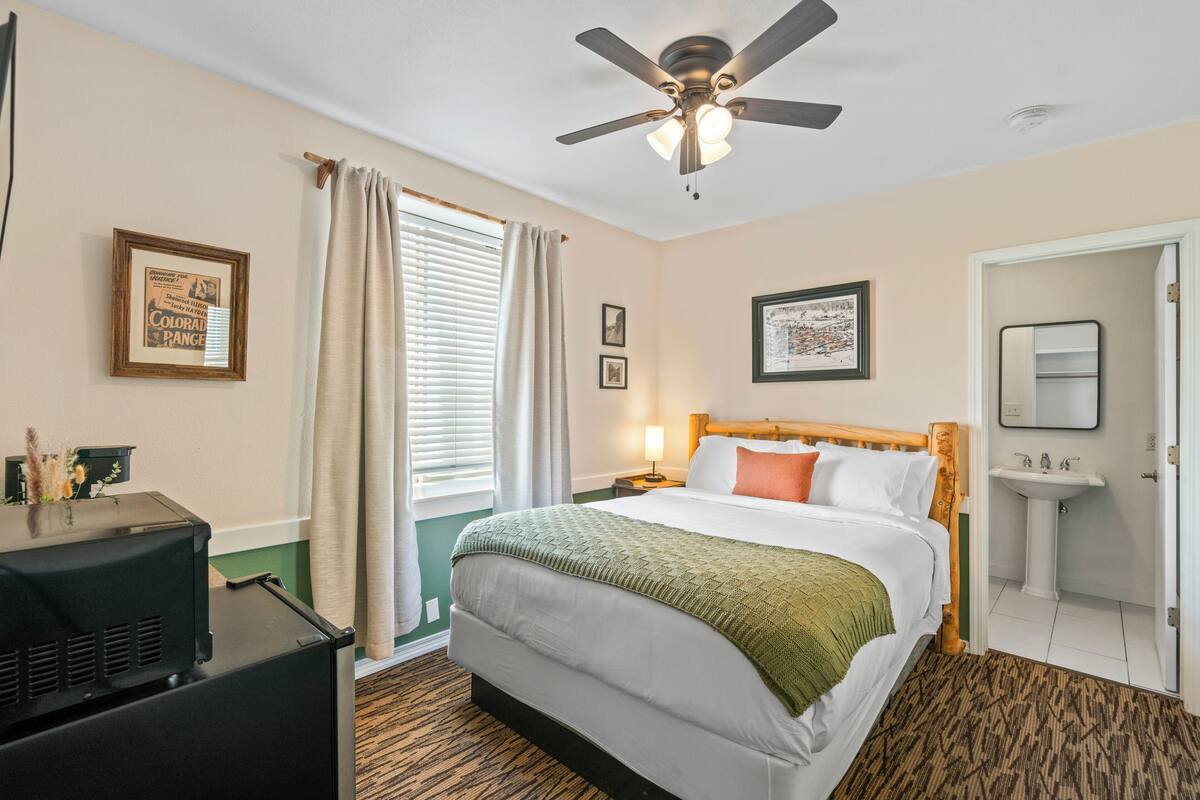 A hotel room with a bed, a ceiling fan, a nightstand, a lamp, a window with curtains, a framed picture, a sink, and a mirror.