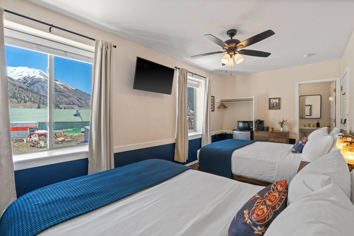 A hotel room with two beds, a ceiling fan, a TV, a desk with a chair, and a window with a view of mountains.