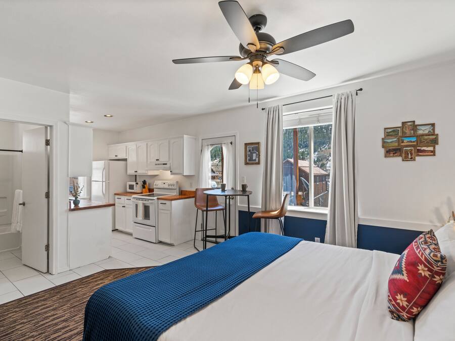 A compact hotel room with a kitchenette, a bed with a blue accent, a ceiling fan, and a view of the outdoors.