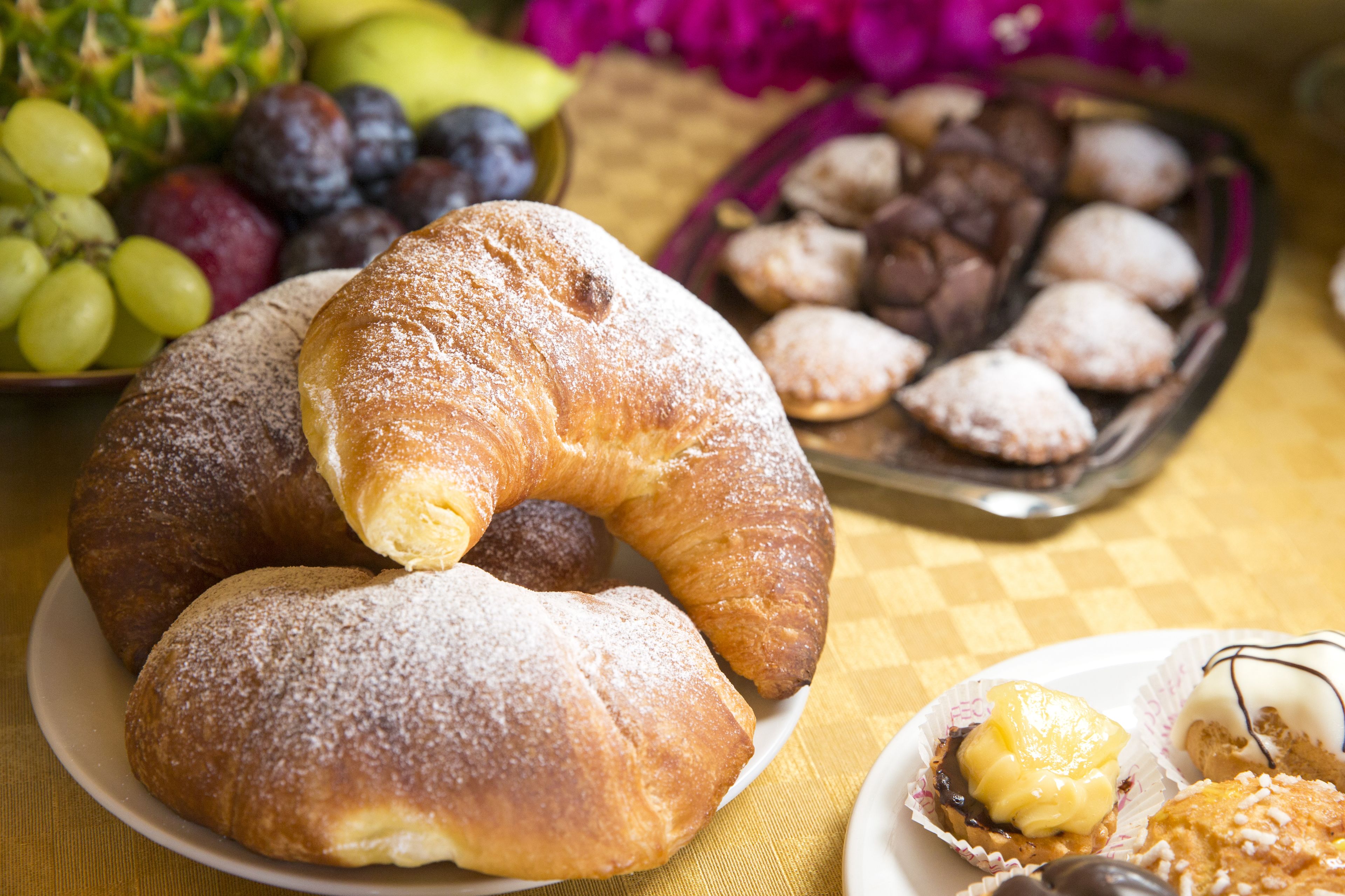 Uma mesa com doces, incluindo croissants e pastéis em copos de papel, e uma tigela de fruta.