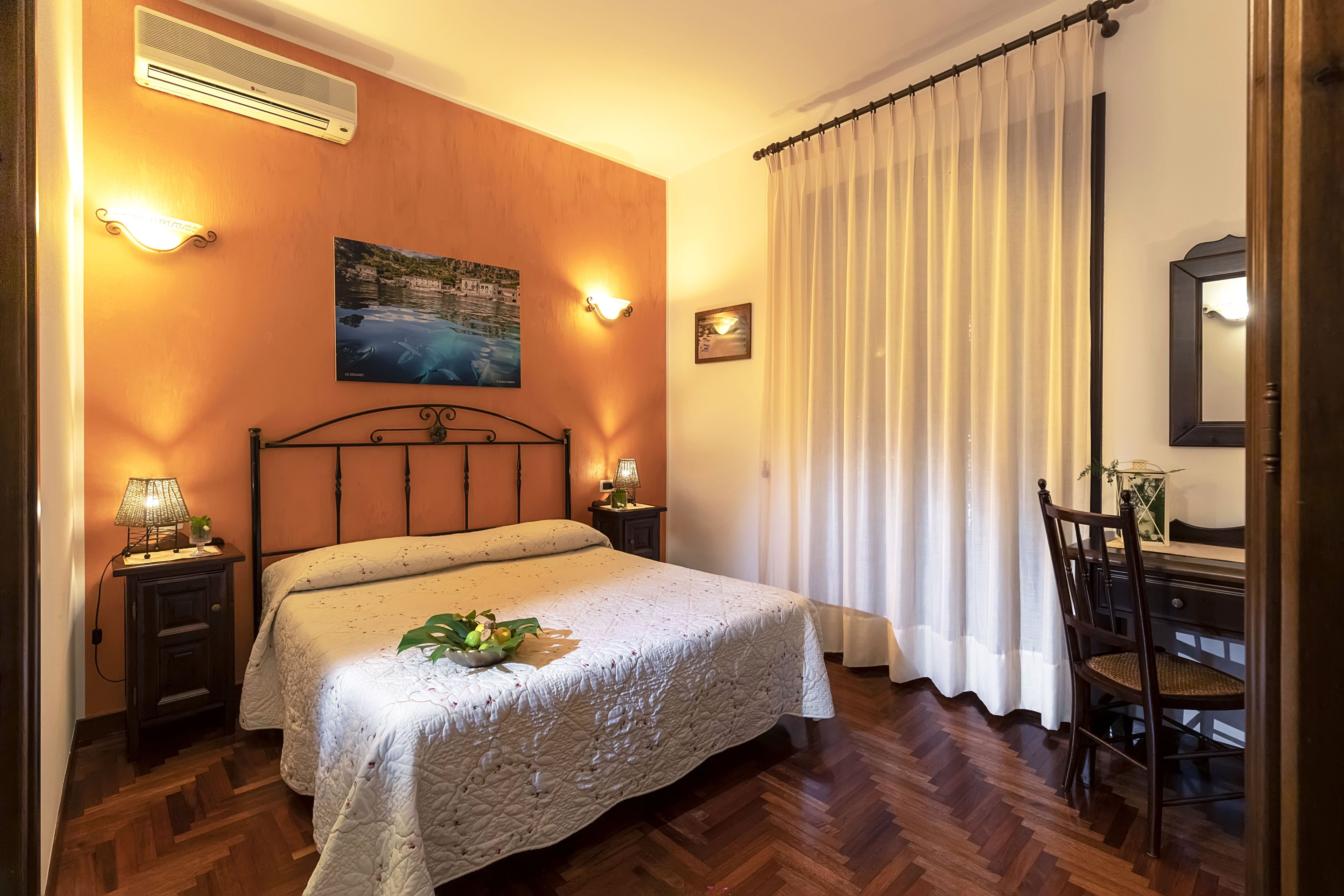 Un dormitorio con cama, lámparas de noche, mesita de noche, una silla, escritorio, espejo y un cuadro en la pared.