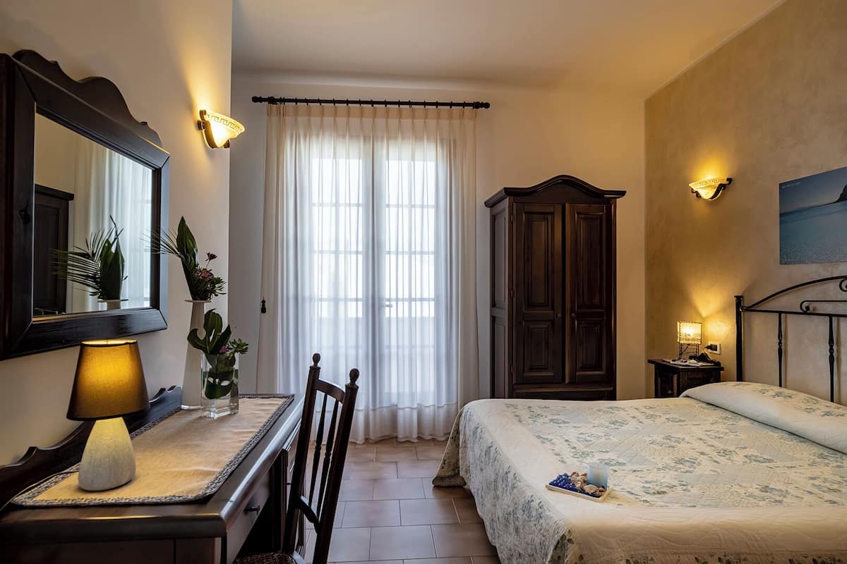 A bedroom with a bed, a desk with a lamp, a wardrobe, and a large window with curtains.