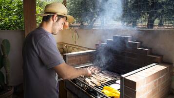 A person grilling meat on an outdoor barbecue with a brick oven and a wire mesh fence in the background.