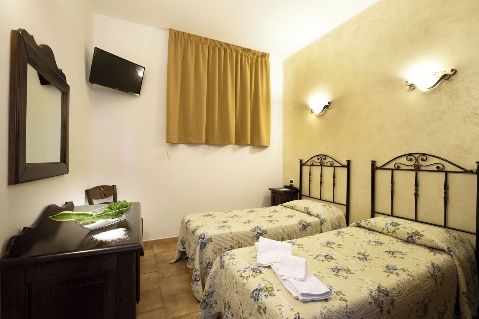A bedroom with two beds, a TV, a mirror, a bedside table, and a window with curtains.