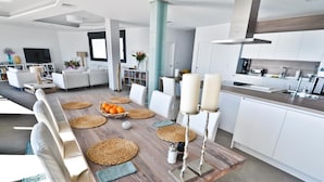 Une cuisine moderne avec des meubles blancs, une table à manger en bois et des sets de table en fibres tressées, ainsi qu’une grande fenêtre.