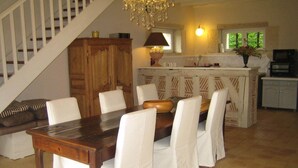 Uma sala de jantar com mesa de madeira e cadeiras brancas, uma escada, um candelabro e uma cozinha ao fundo.