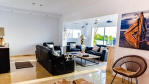 Ein modernes Wohnzimmer mit einer schwarzen Ledersofa, einem Holztisch und einem braunen Fellteppich. Ein Korbsessel und ein Gemälde eines Bootes sind an der Wand zu sehen.