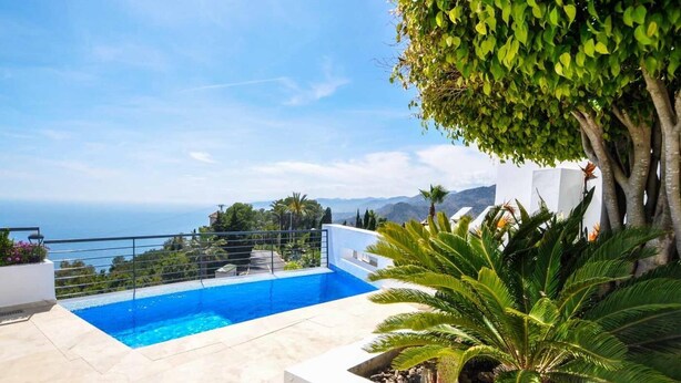 Ein Pool mit freiem Blick auf das Meer, umgeben von einem weißen Gebäude und üppiger Vegetation.