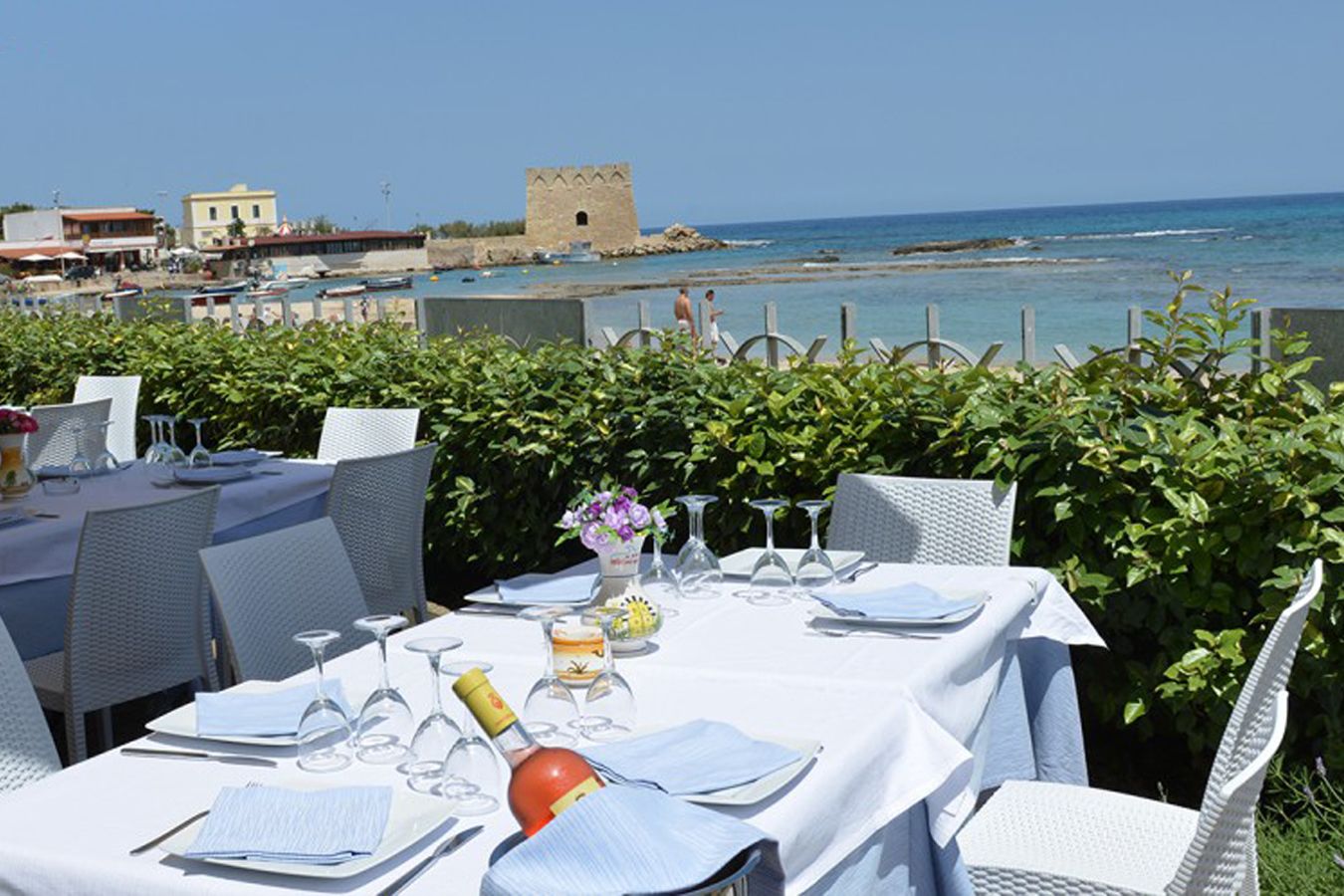 Un área de comedor al aire libre con mesas dispuestas para una comida, con vista al mar y un edificio histórico al fondo.