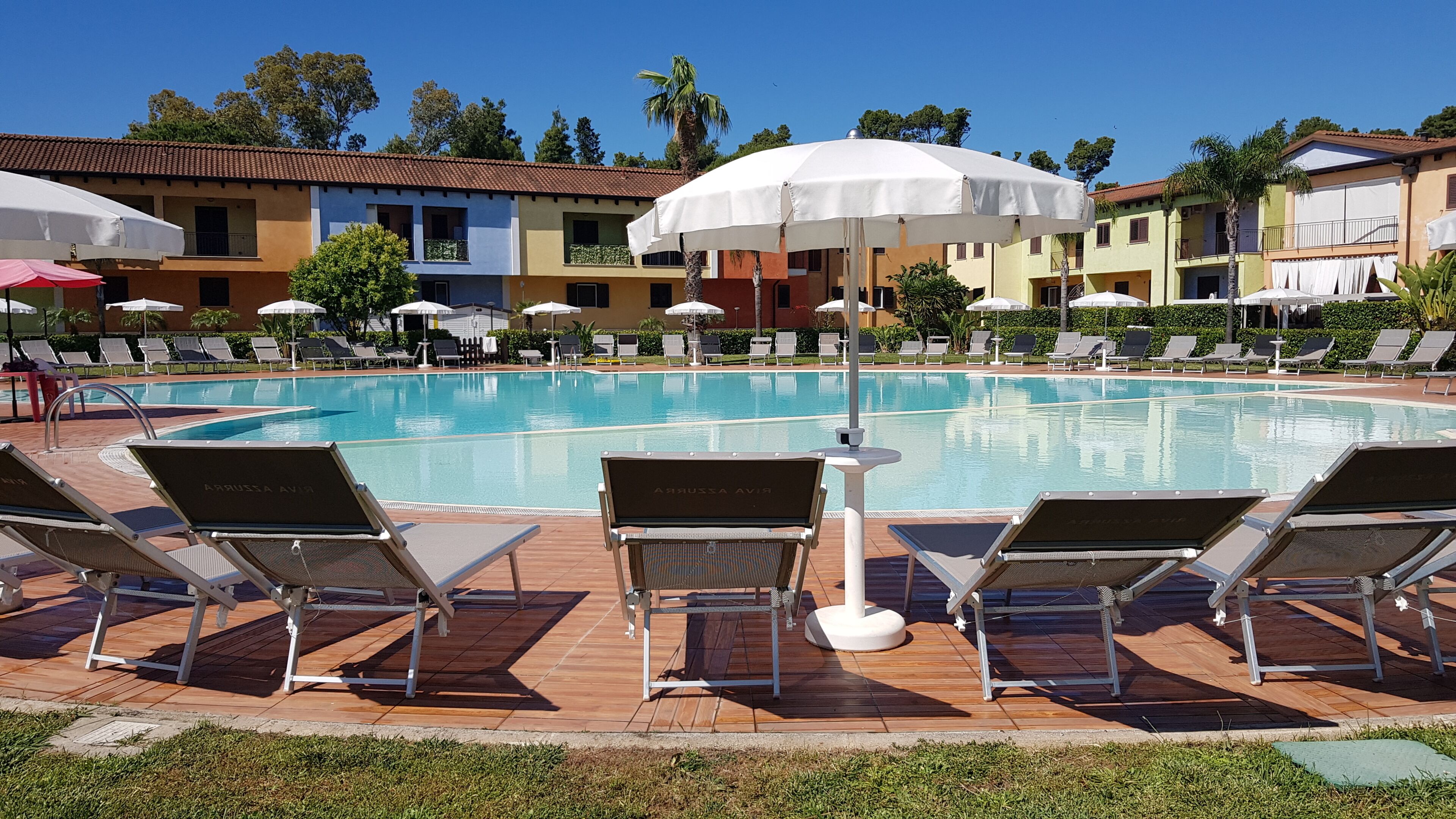 Uma área à beira da piscina com espreguiçadeiras e guarda-sóis, piscina azul cristalina e edifícios ao fundo.