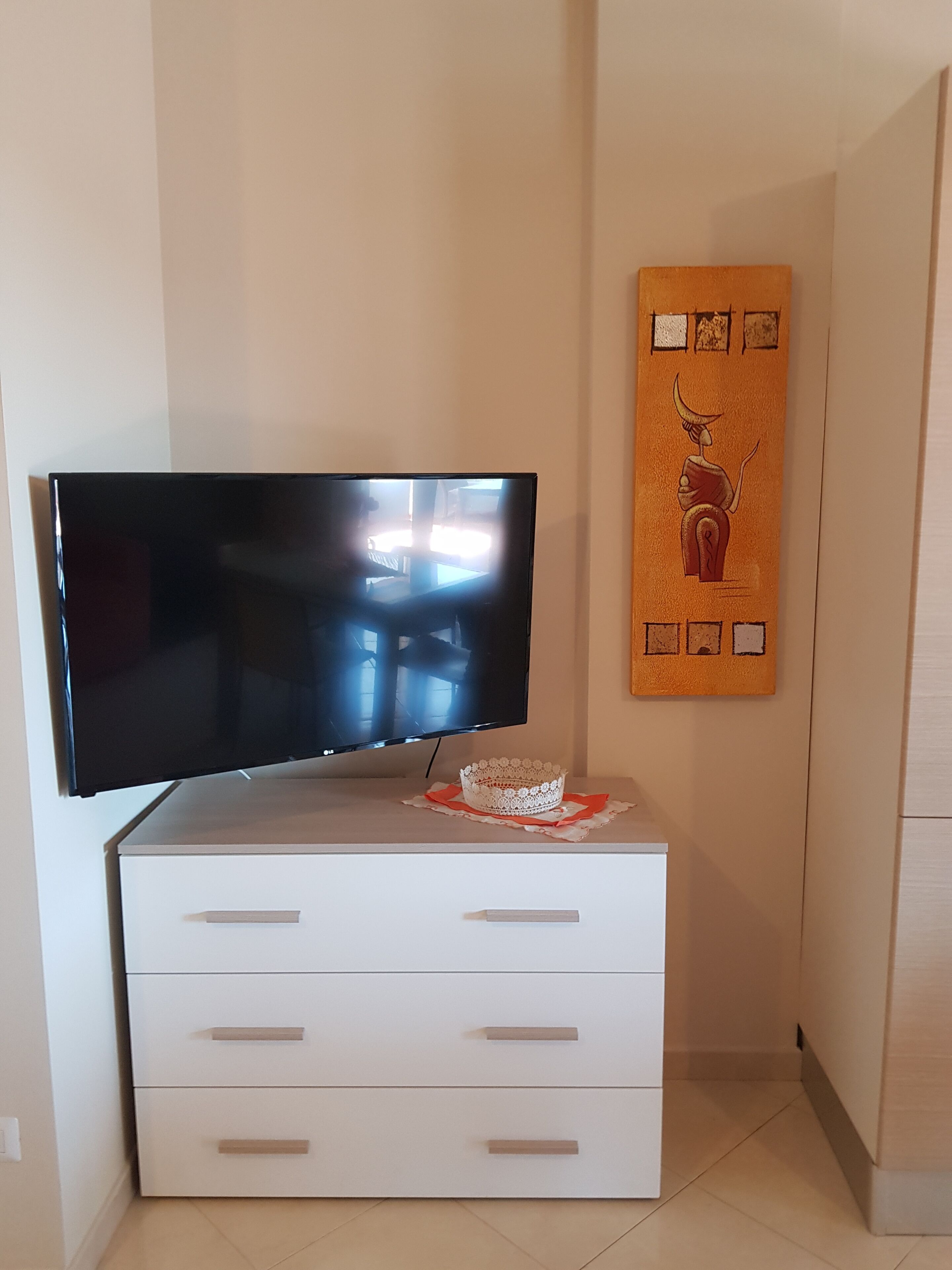 Ein Flachbildfernseher an einer Kommode mit Schubladen, ein dekorativer Gegenstand darauf und ein gerahmtes Kunstwerk an der Wand.